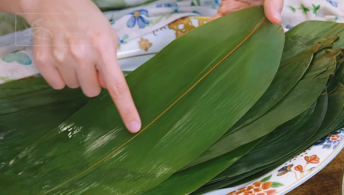 Wrap zongzi: Place two bamboo leaves together (rough side out, smooth side in), forming a cone shape. Add glutinous rice, pork, meantại, and more rice, packing tightly. Fold and tie with kitchen twine.